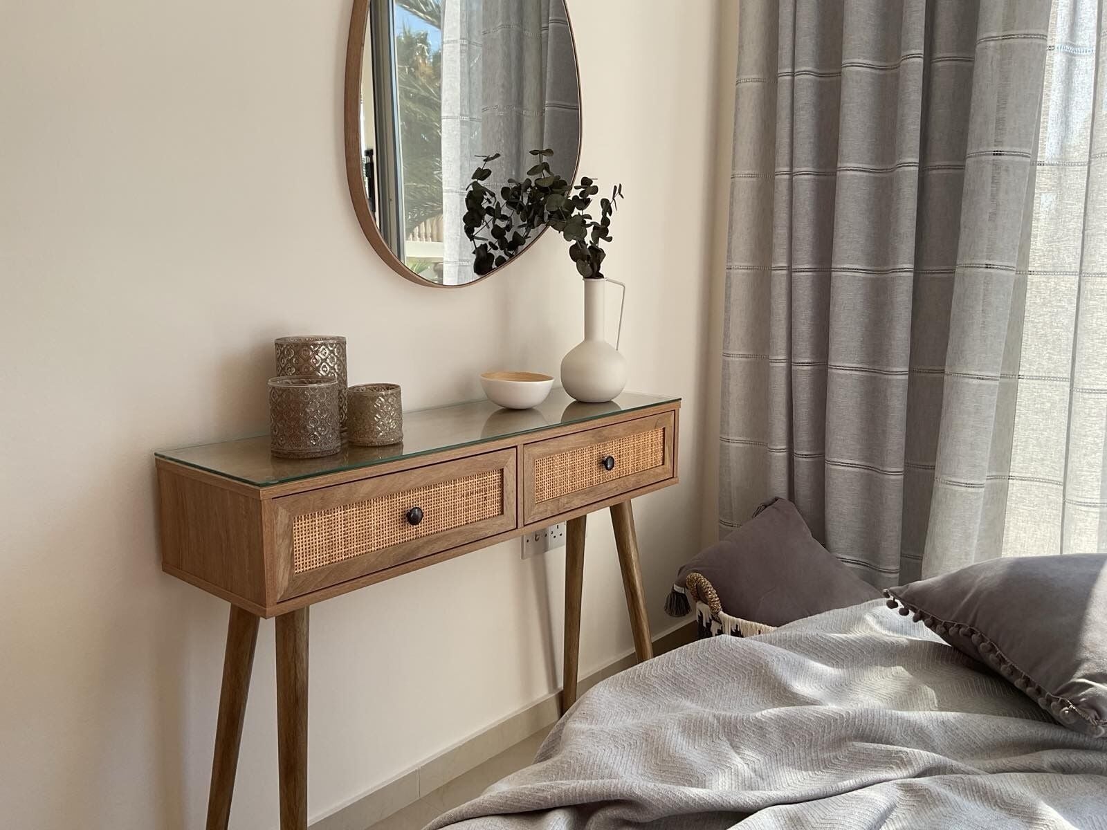 Een slaapkamer met een bed, een nachtkastje met decoratieve objecten, een spiegel en een raam met gordijnen.