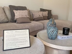 Ein gemütlicher Wohnbereich mit einer Couch, dekorativen Kissen, einer blauen Vase, einer Kerze und einer begrüßenden Nachricht im Bilderrahmen.