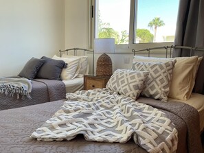 Ein Schlafzimmer mit zwei Betten, einem Fenster mit Blick auf Palmen und einer Nachttischlampe.