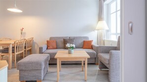 Ein Wohnzimmer mit einer Couch, zwei Sesseln, einem Couchtisch, einem Esstisch mit Stühlen und einer Vase mit Blumen.