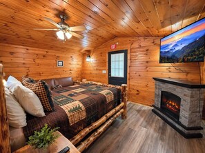 Ein gemütliches Blockhaus-Zimmer mit einem Bett, einem Kamin, einem Fernseher und einem Deckenventilator.