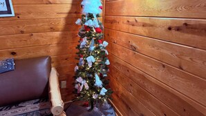 Interior de una cabaña de madera con un árbol de Navidad decorado, un sofá de cuero marrón y una pared de madera.
