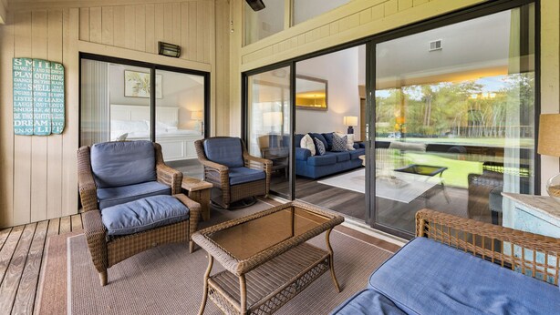 Una habitación amplia con muebles de mimbre, ventilador de techo y ventanas grandes con vistas al exterior.