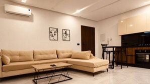 Un salon moderne avec un canapé d’angle beige, une table basse et un coin cuisine doté d’une cuisinière noire et de meubles de rangement en bois.