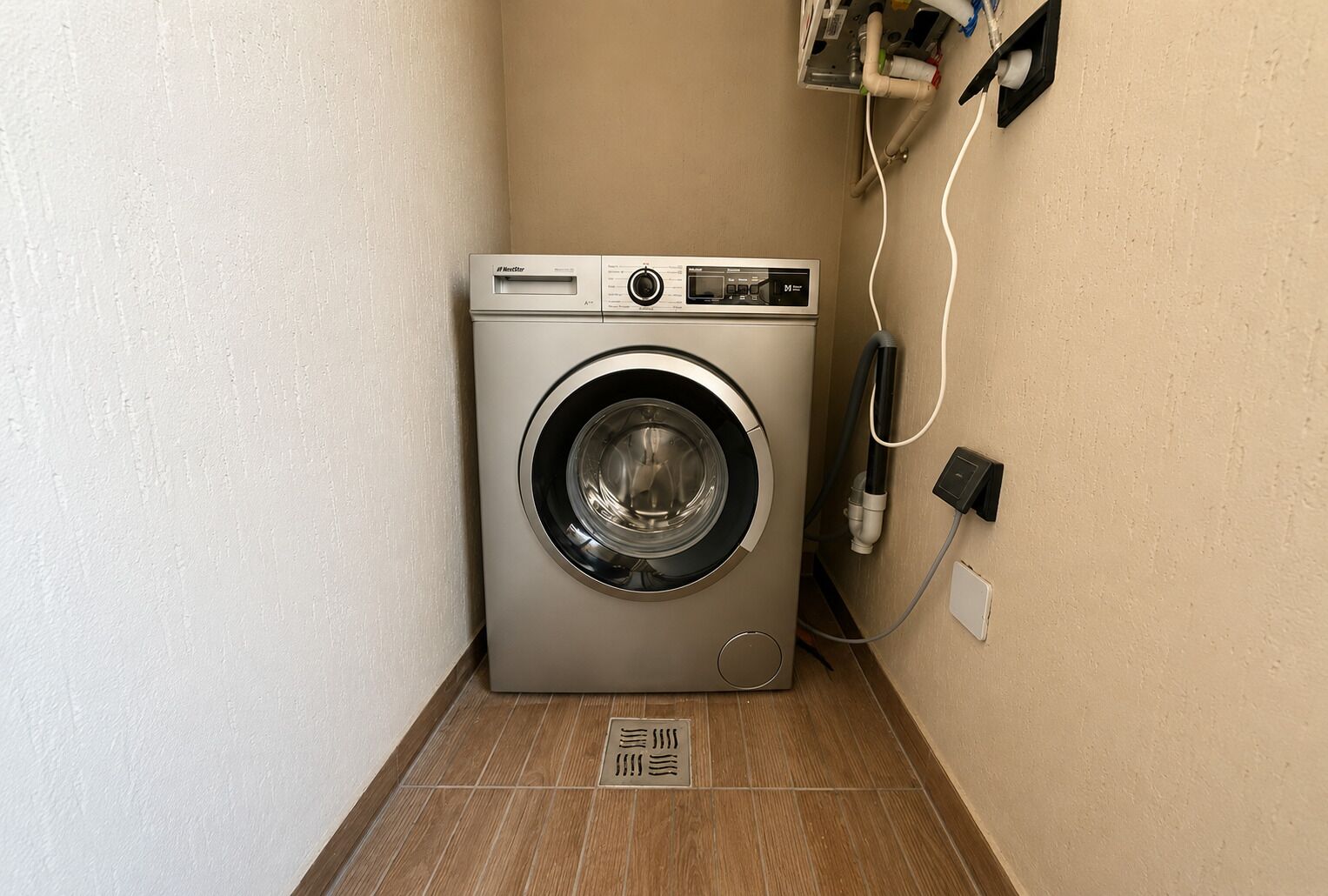 Een compacte, moderne wasmachine in een hoek van een kamer met een betegelde vloer en een effen muur.