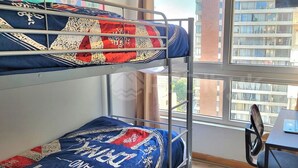 Un lit superposé avec une literie à motifs, un bureau avec un ordinateur portable et un ventilateur, et une fenêtre donnant sur des immeubles.