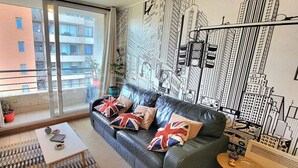 Un salon moderne avec un canapé, des coussins, une table basse et une fresque murale représentant un paysage urbain.