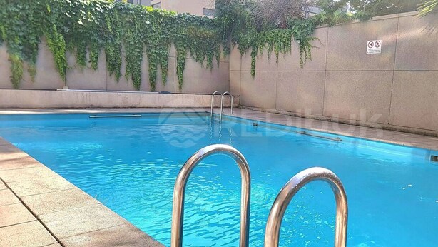 Une piscine avec des échelles en métal, entourée d’une terrasse carrelée et d’un petit muret en pierre agrémenté de verdure.