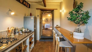 A narrow kitchen with a refrigerator, stove, and wooden cabinets. A dining area with a table and chairs is visible.