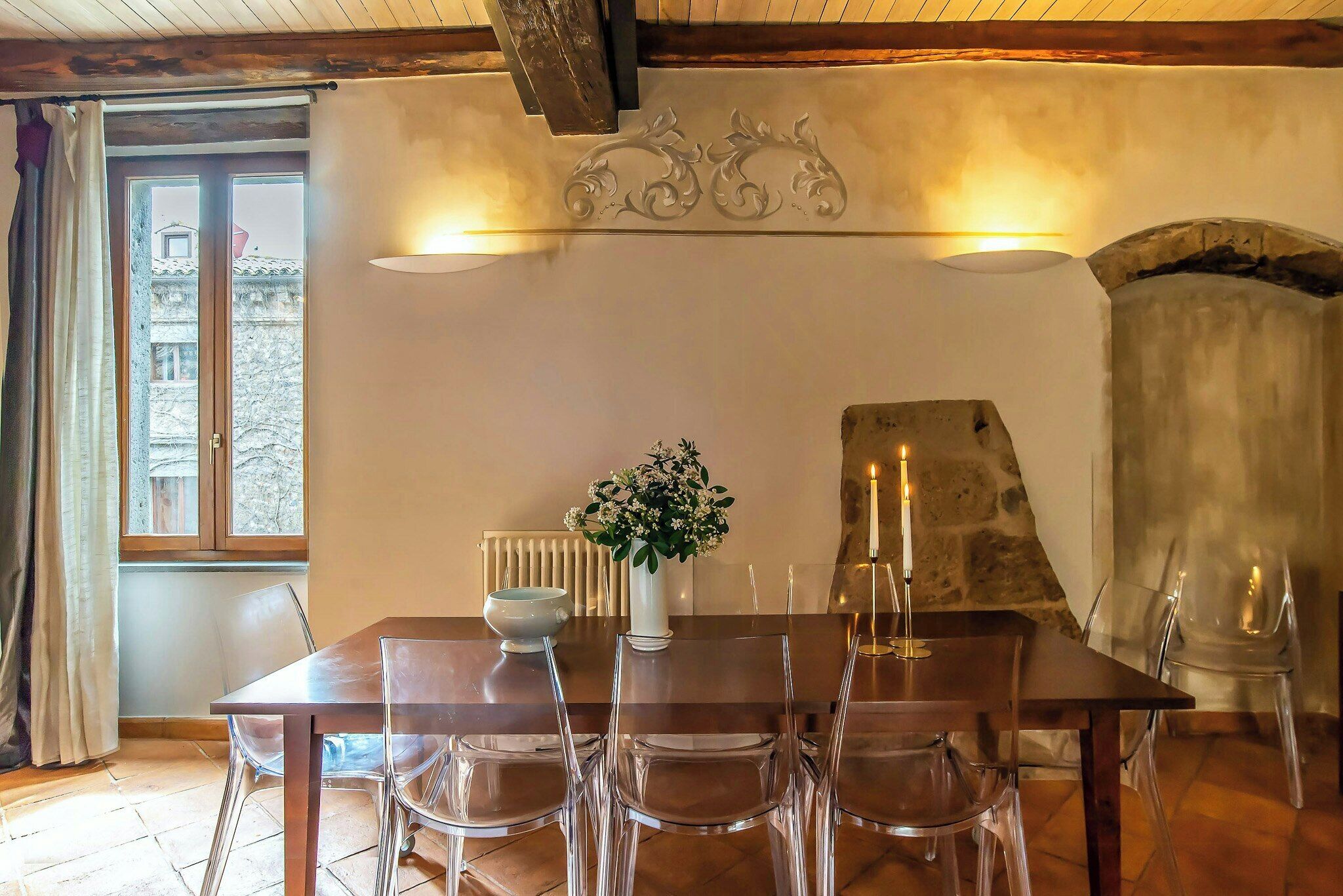 A dining area with a wooden table, clear chairs, a vase with flowers, and a stone fireplace.