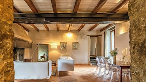 A rustic interior with exposed wooden beams, a fireplace, a dining area with clear chairs, and a white sofa.