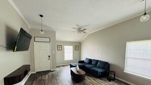 Ein Wohnzimmer mit dunklem Holzboden, einem dunkelblauen Sofa, einem Couchtisch, einem an der Wand befestigten Fernseher, einem Deckenventilator und einem Fenster mit Jalousien.