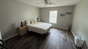 Ein Schlafzimmer mit einem Bett, Nachttischen, einem Deckenventilator, einem Fenster mit Jalousien, einem kleinen Regal und einem Papierkorb.