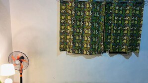 Una habitación con una cama, un ventilador de pie y una ventana con cortinas estampadas.