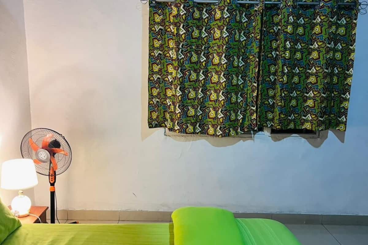A room with a bed, a standing fan, and a window with patterned curtains.
