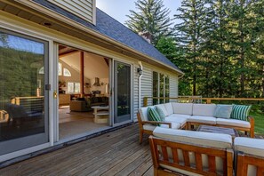 A wooden deck with a sofa and chairs, a sliding glass door leading to a kitchen, and a view of a forested area.