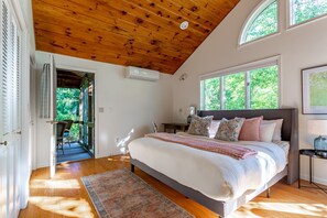 A bedroom with a wooden ceiling, a large bed, a nightstand, a window with a view of greenery, and a door leading to a balcony.