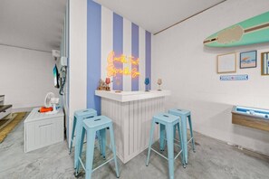 Eine Bar mit einer Neonreklame, zwei Barhocker, einem Ventilator und einer Surfbrett-Dekoration.