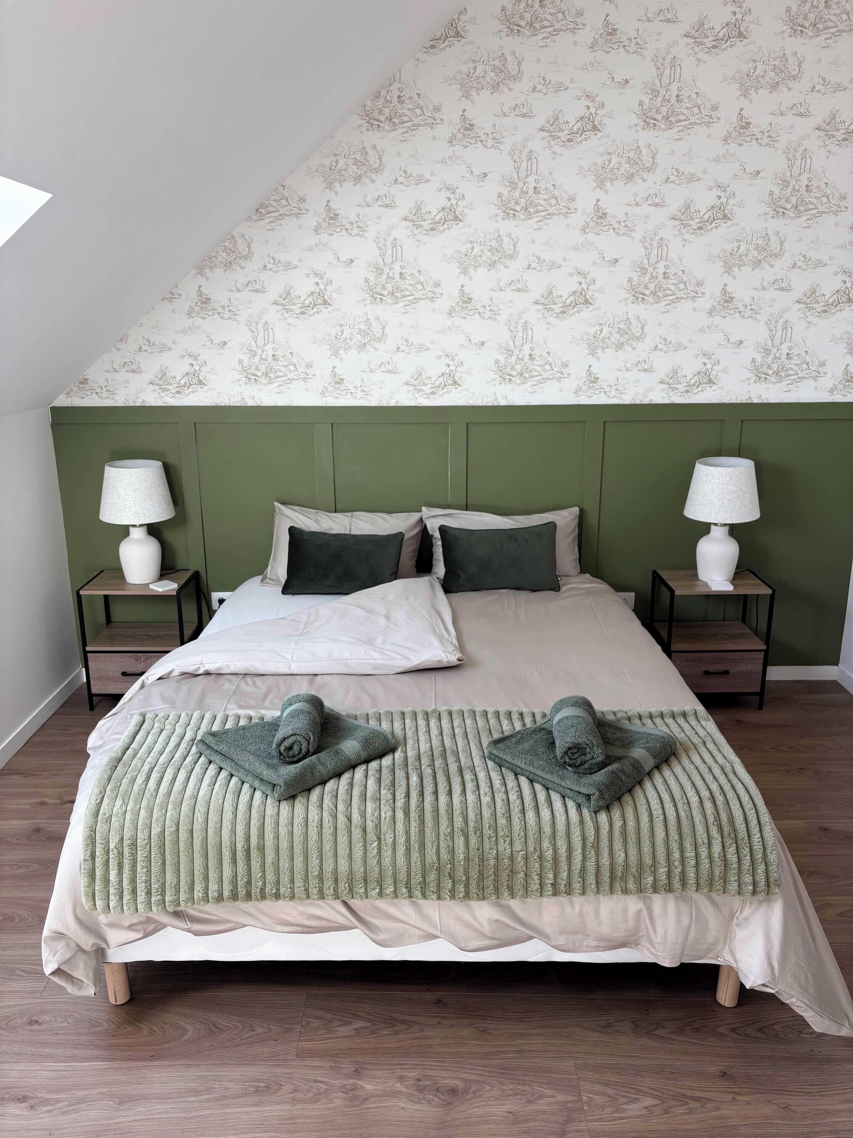 A bedroom with a green headboard, two bedside tables with lamps, a bed with a green blanket, and a patterned wallpaper.