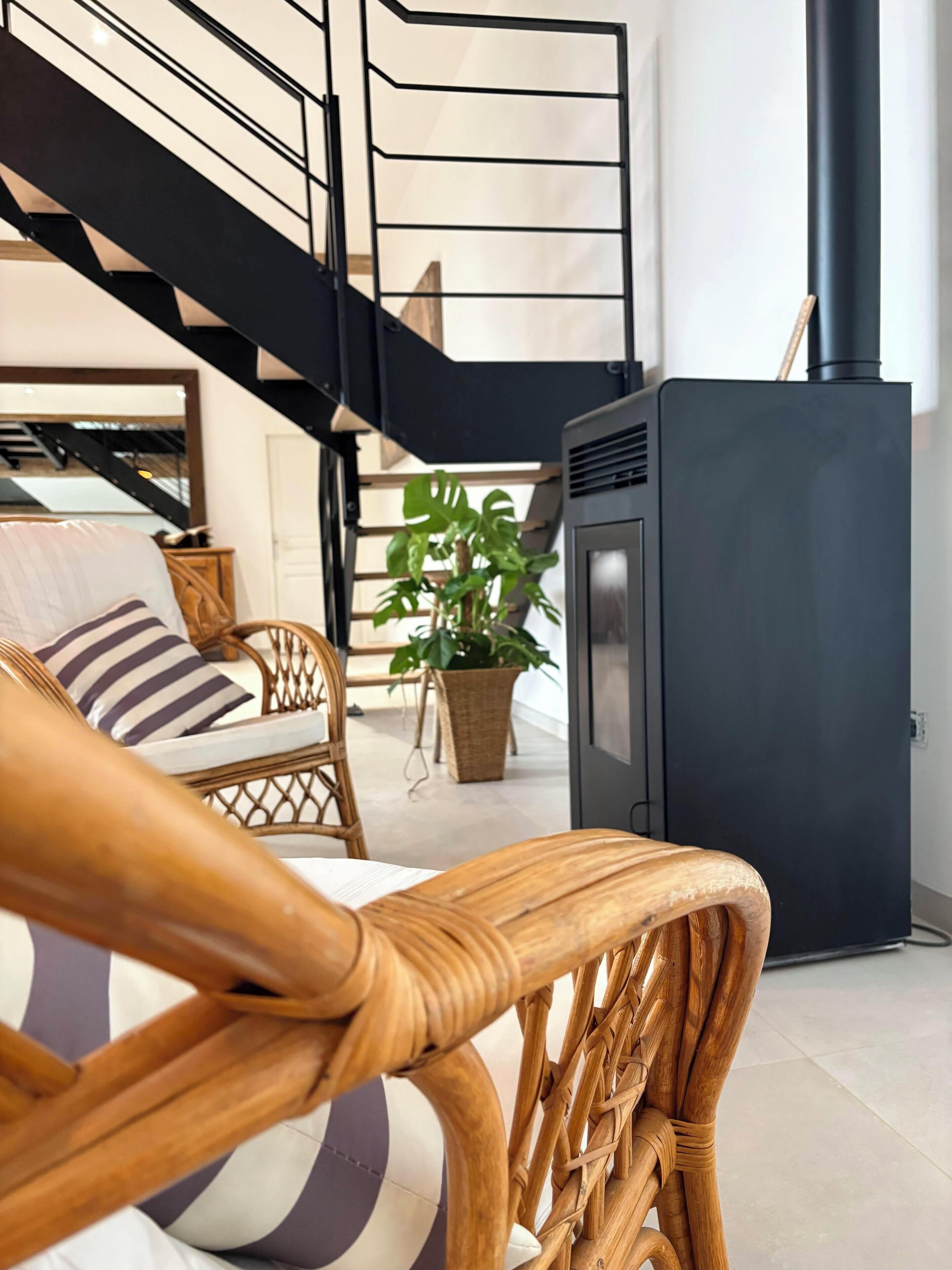A modern living room with a sofa, a wicker chair, a potted plant, a black fireplace, and a staircase.