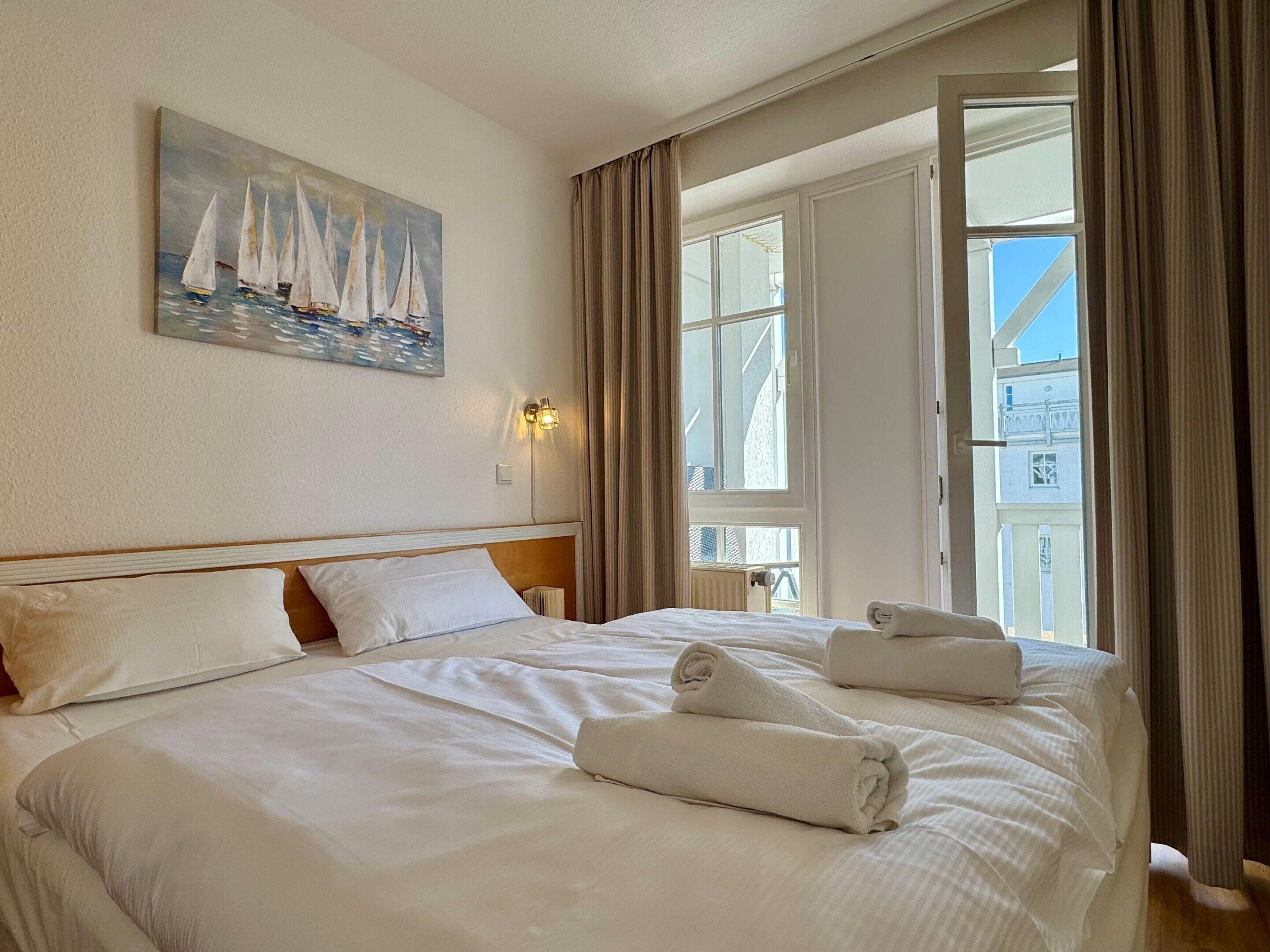 Ett hotellrum med en stor säng, en målning som föreställer segelbåtar, ett fönster med gardiner och utsikt över byggnader utanför.