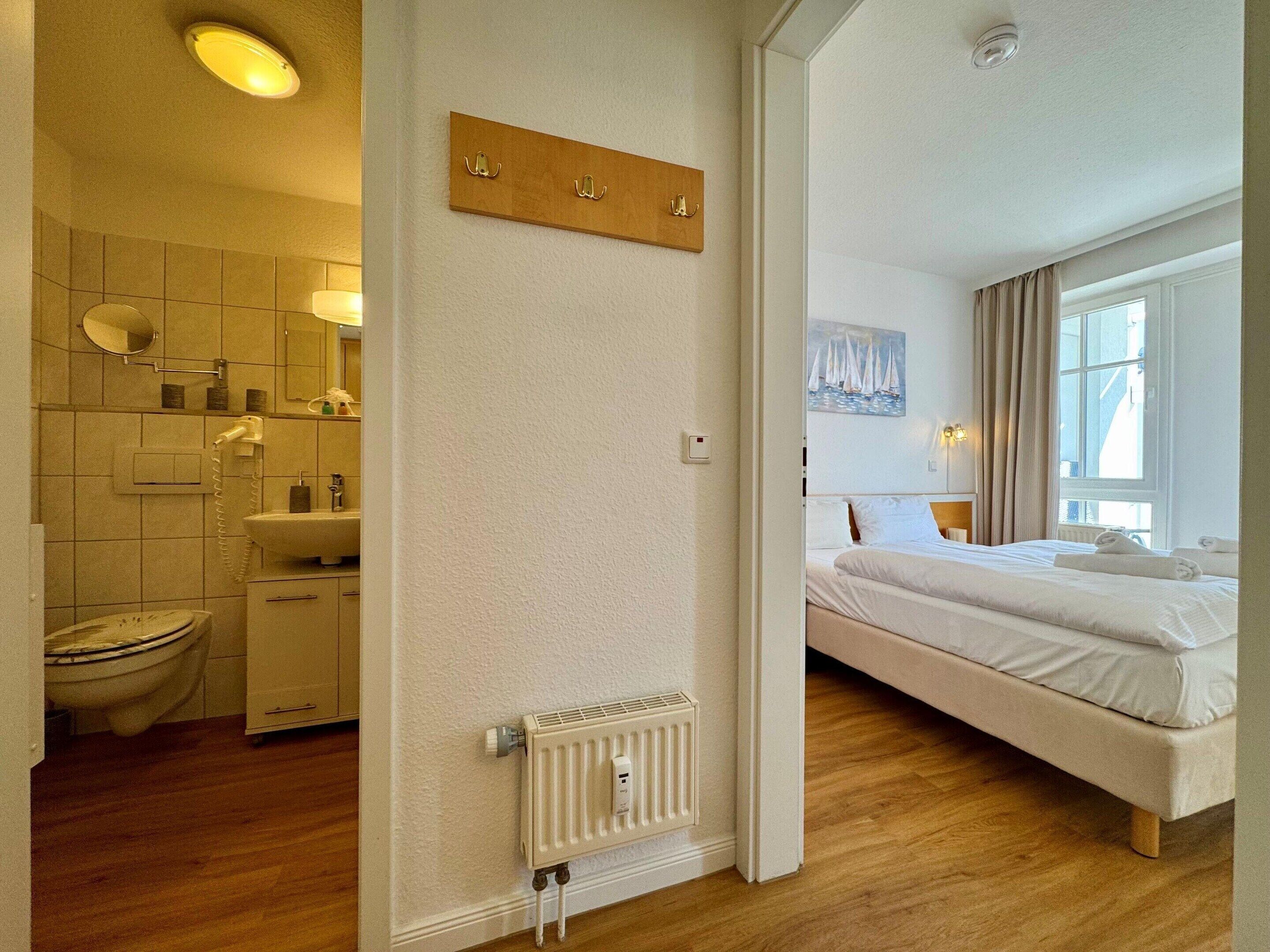 Habitación de hotel con suelo de madera, cama con ropa de cama blanca, radiador, baño con inodoro, lavabo y espejo, y un cuadro en la pared.