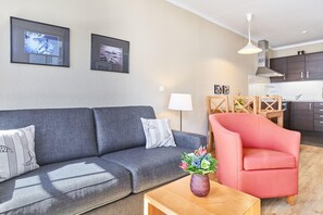 Ein modernes Wohnzimmer mit einem grauen Sofa, einem roten Sessel und einem Couchtisch aus Holz. An den Wänden hängen Bilderrahmen und auf einem Beistelltisch steht eine Lampe.