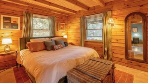 A wooden cabin bedroom with a bed, pillows, a nightstand, a lamp, and a window with curtains. - The Oak & Oar Camphouse | Family Lake Retreat (Logan)