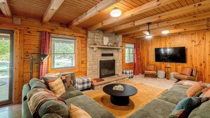 A log cabin interior with a stone fireplace, a large flat-screen TV, and a cozy seating area with a sofa and ottomans. - The Oak & Oar Camphouse | Family Lake Retreat (Logan)