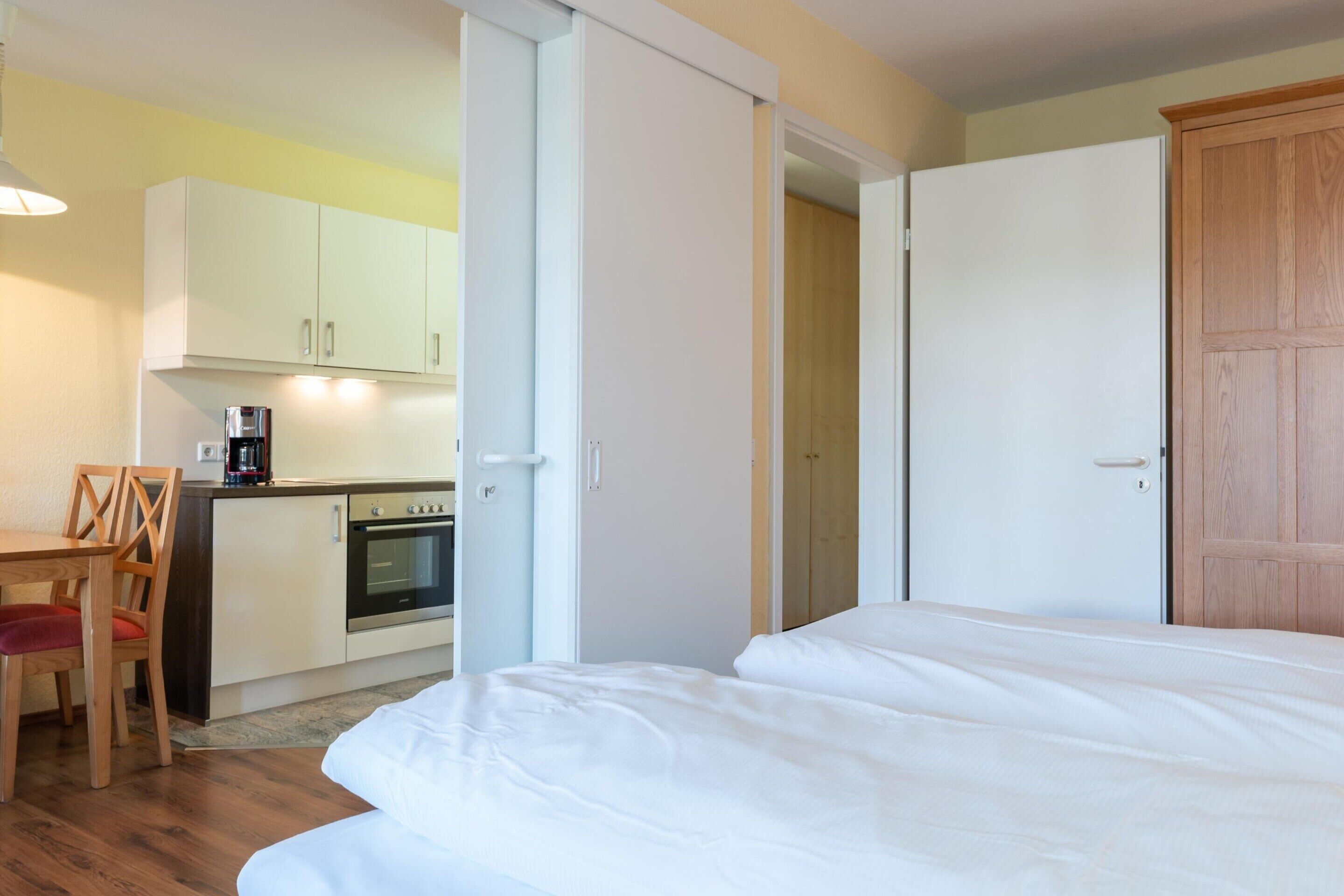 A bedroom with a bed, a wooden door, and a kitchenette with cabinets and a coffee maker.