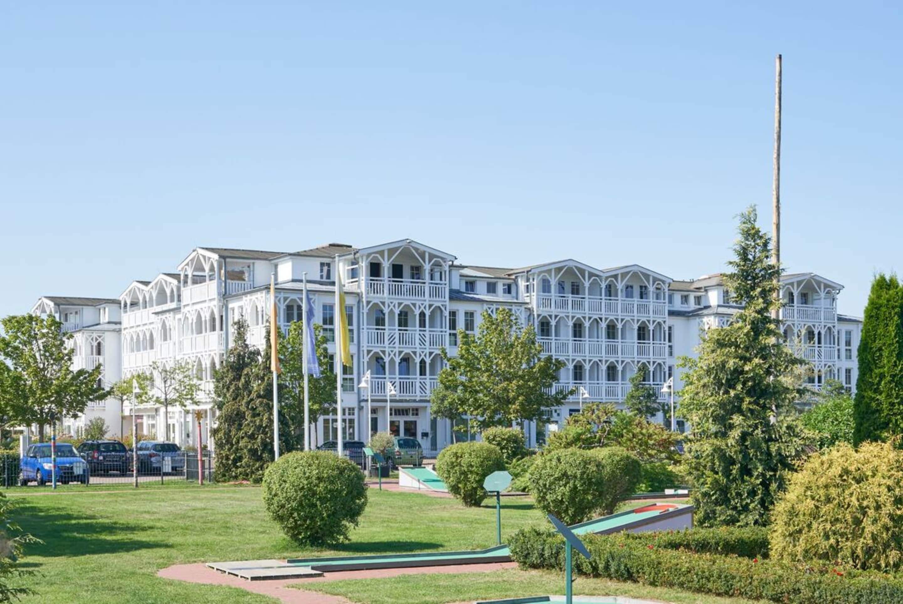 Ein großes, weißes, mehrgeschossiges Gebäude mit symmetrischer Fassade und Balkonen, umgeben von gepflegter Bepflanzung und blauem Himmel.