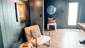 A room with a chair, a framed picture, a round mirror, and a door with a view.