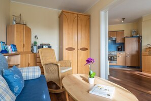Ein Wohnzimmer mit einer Couch, einem Korbstuhl, einem Holzschrank und einem kleinen Tisch mit einem Buch und einer Blume.