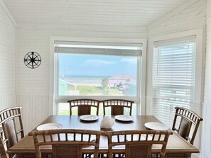 Ein Essbereich mit Tisch und Stühlen, einer Kompass-Wanddekoration und Blick auf den Strand und Häuser durch das Fenster.
