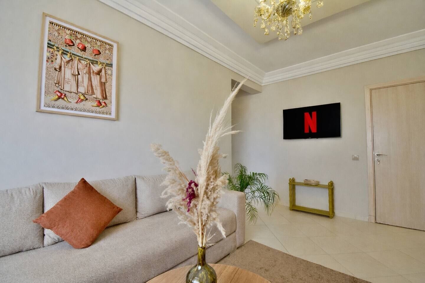 En stue med en sofa, en vase med tørrede planter, et indrammet billede, et Netflix skilt og en dør.