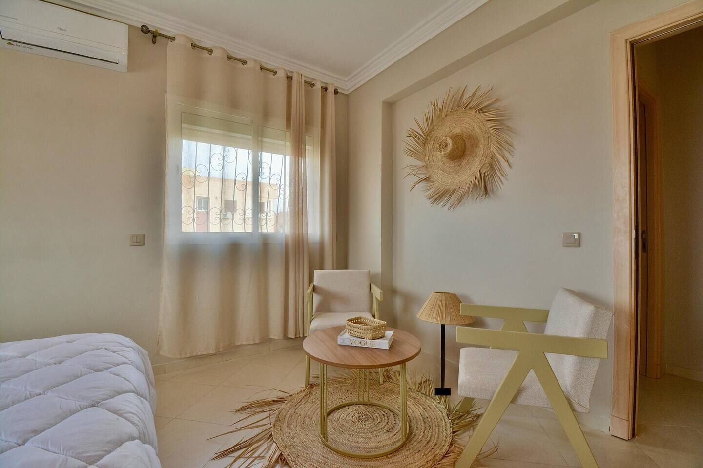 A room with a bed, a chair, a small table with a book, a lamp, and a wall-mounted decorative item.