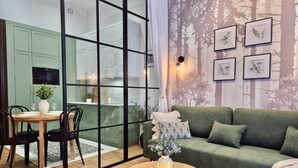 Ein modernes Wohnzimmer mit einem grünen Sofa, einem Esstisch mit Stühlen und einer durch eine Glastrennwand sichtbaren Küche.