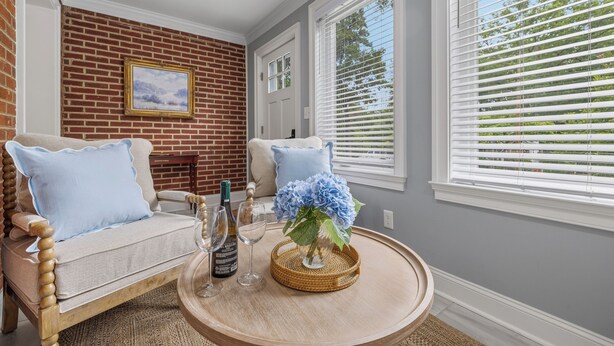 A cozy room with a brick wall, two sofas with blue pillows, a round wooden table with a vase and wine glasses, and large windows with blinds.