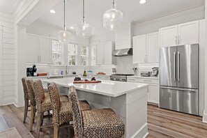 A modern kitchen with white cabinetry, a stainless steel refrigerator, and a central island with bar stools. - 30A Family Retreat | Pool, Tram & Outdoor Living (Inlet Beach)
