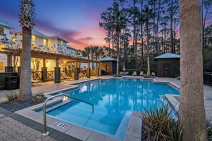 A swimming pool with a wooden deck, surrounded by palm trees and a building with a covered patio. - 30A Family Retreat | Pool, Tram & Outdoor Living (Inlet Beach)