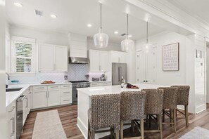 A modern kitchen with white cabinetry, a central island, bar stools, pendant lighting, and a backsplash with a geometric pattern. - 30A Family Retreat | Pool, Tram & Outdoor Living (Inlet Beach)