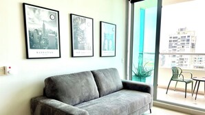 Una sala moderna con un sofá gris, cuadros con vistas de la ciudad en la pared, una planta en maceta y vistas de edificios a través de un ventanal grande.