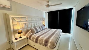 Un dormitorio moderno con cabecera capitonada, una cama grande con ropa de cama a rayas, una mesita de noche con lámpara, un ventilador de techo y un televisor montado en la pared.