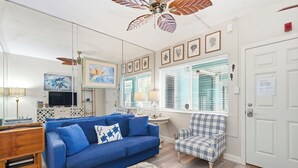 A living room with a blue sofa, a plaid armchair, a ceiling fan, and framed artwork on the walls. - Turquoise Dream  One bedroom Condo On Pass a Grille Beach (St. Pete Beach)