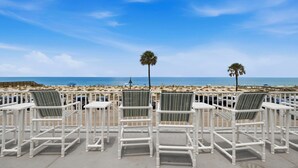 A beachside patio with white chairs and tables, palm trees, and a view of the ocean. - Turquoise Dream  One bedroom Condo On Pass a Grille Beach (St. Pete Beach)