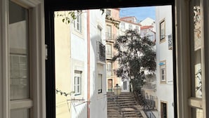 Vista a través de una ventana abierta de una calle estrecha flanqueada por edificios, una escalera y un árbol.