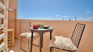 Ein Balkon mit zwei Stühlen, einem kleinen Tisch und einer Schale mit Essen, vor einem wolkenlosen, blauen Himmel.