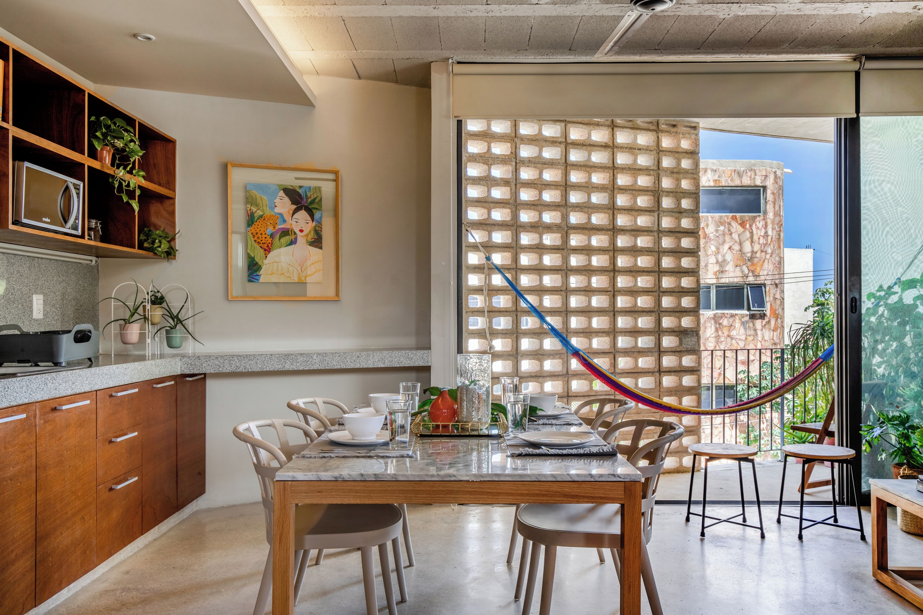 A dining area with a table set for four, surrounded by chairs, a kitchen with wooden cabinets, and a large window with a view of buildings.