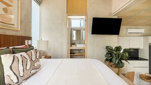 Habitación de hotel con cama, televisor, planta, kitchenette y baño con espejo y lavamanos.