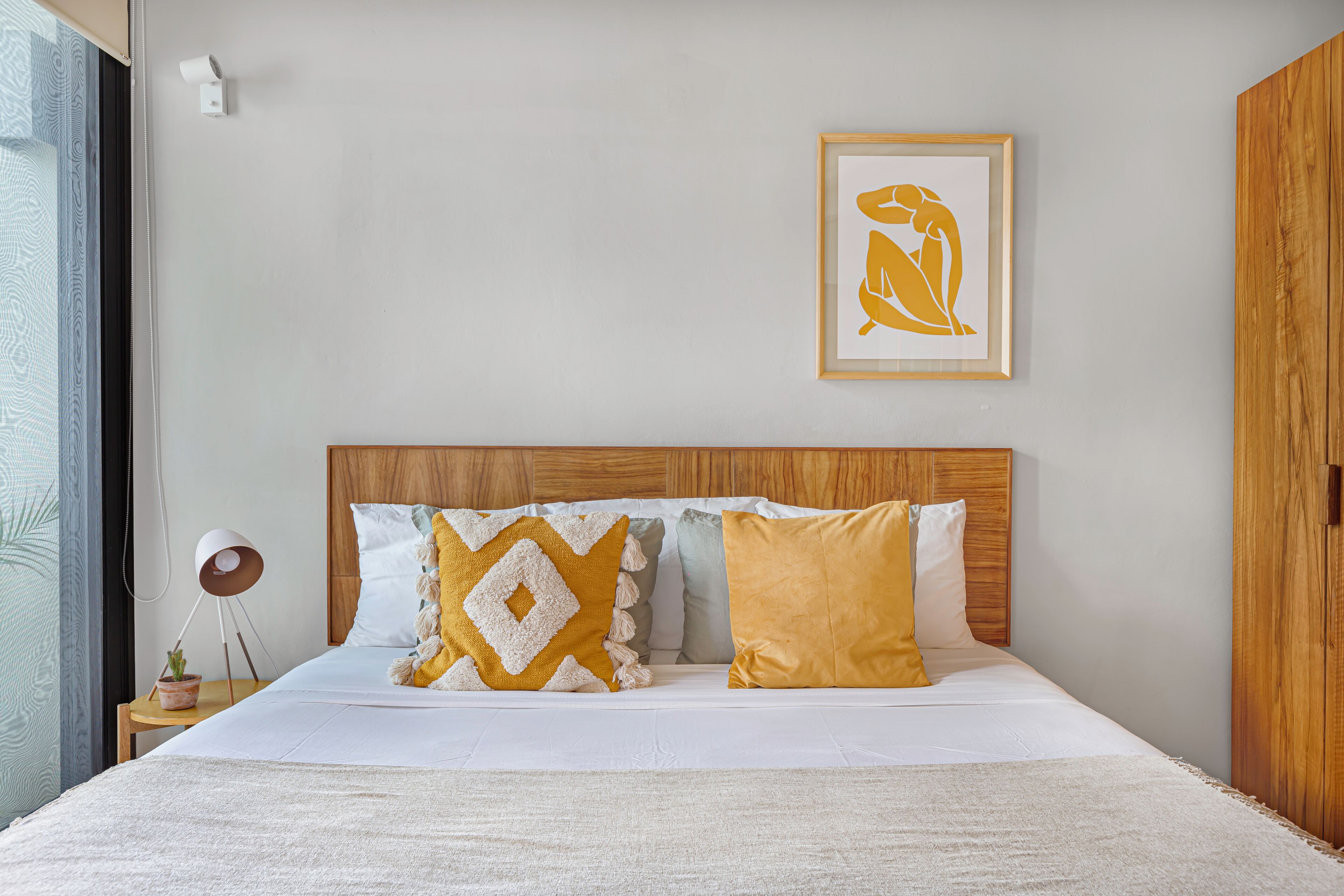 A neatly arranged bedroom with a bed, pillows, a framed artwork, and a small table with a plant.